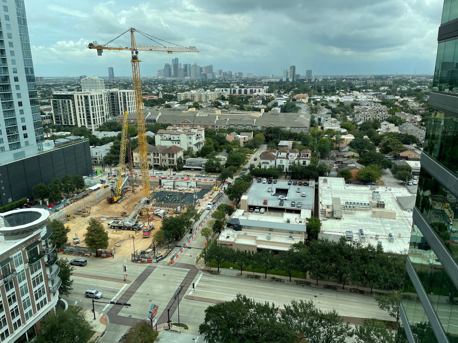 An unannotated aerial photograph on an overcast day, looking east across a city. A yellow tower crane and a deep excavation occupy the left foreground; a two-level parking deck and a large flat-roofed building with rooftop refrigeration units sit centre right; a wide boulevard crosses the bottom of the frame. Beyond them, blocks of townhomes, and on the horizon a cluster of tall towers in haze.